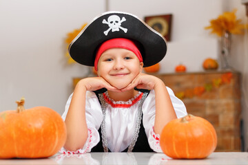 Celebrating Halloween at home. A little girl dressed as a pirate sits at a table with pumpkins. - obrazy, fototapety, plakaty