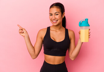 Young Venezuelan woman drinking a protein shake isolated on pink background smiling and pointing...