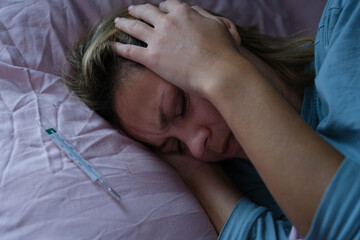 Woman lying in bed and holding sore head with hands near mercury thermometer