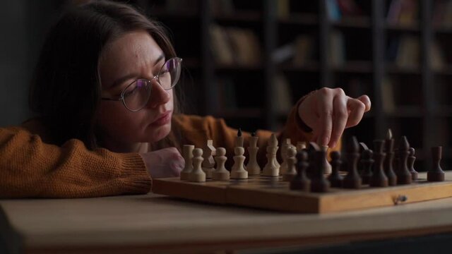Close-up face of pensive female chess player performing move with chess pieces on old wooden board. Thoughtful young woman wearing glasses playing chess alone at home on blurred background of bookcase