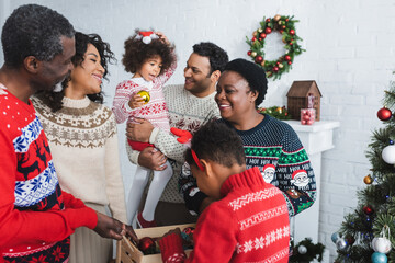 boy holding wooden box with christmas balls near cheerful african american family in living room