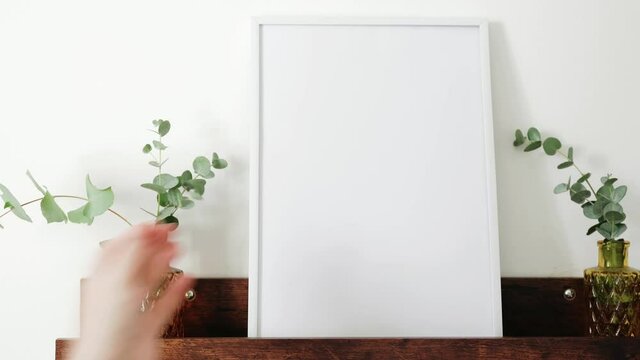 Female hand placing white picture frame on brown shelf against white wall. Mock up. Copy space. 4K UHD footage
