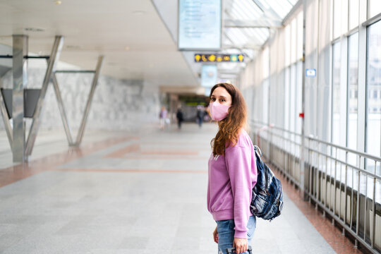 Sad young woman in a protective mask with backpack stands at the train station or in the airport. Copy space.