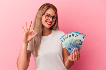 Young russian woman holding bills isolated on pink background cheerful and confident showing ok gesture.