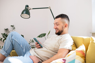 Obraz premium Happy positive man text messaging on cellphone while resting on couch in living room. Guy lying on couch reading messages on mobile phone.