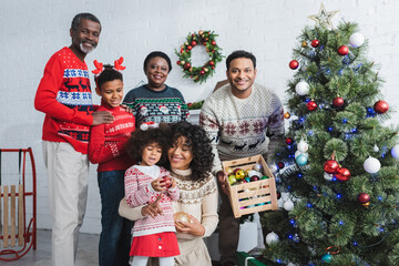 man holding wooden box with decorative baubles near christmas tree and african american family