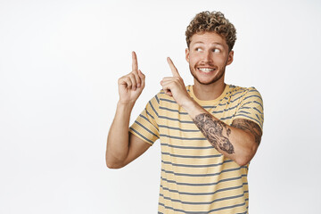 Cheerful young blond man showing advertisement, pointing fingers at upper left corner and smiling excited, recommending store sale, white background