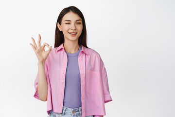 Smiling young woman winking, showing okay, OK sign, assure everything good, making compliment,...