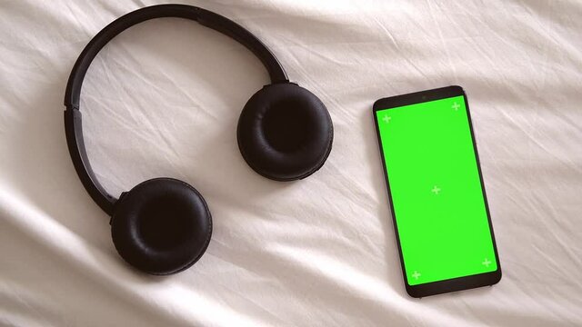 The phone with a green screen lies on a white bed with headphones. Chroma key phone background for music listening application. Music app
