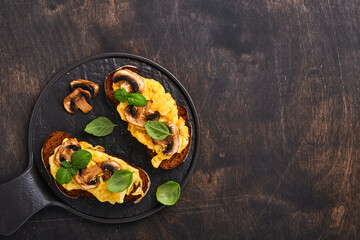 Scrambled eggs with fried mushrooms and basil on bread on black table background. Homemade breakfast or brunch meal - scrambled eggs and mushrooms sandwiches. Top view with copy space