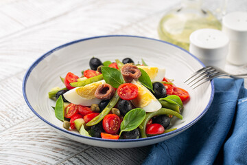 Close-up of mediterranean salad with anchovy, green beans, tomatoes, eggs, black olives, capers on white plate and white wooden background. Healthy food, no carb diet concept.
