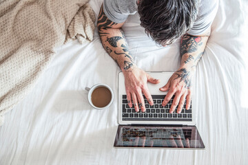 cropped view of tattooed man typing on laptop in bed with coffee cup and laptops