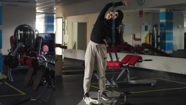 Wide shot positive confident Middle Eastern woman in hijab stretching neck and hand muscles smiling standing in gym. Portrait of beautiful fit slim sportswoman warming up indoors training