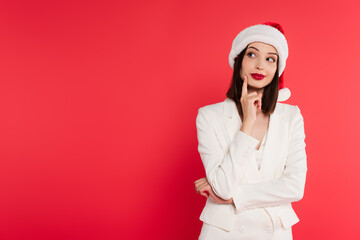 Pensive woman in santa hat and white jacket looking away isolated on red