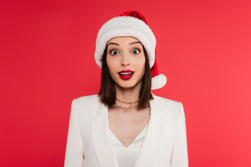 Amazed woman in jacket and santa hat looking at camera isolated on red