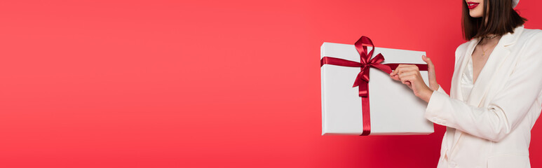 Cropped view of young woman in white jacket holding gift with bow isolated on red, banner