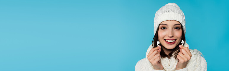 Young woman in warm clothes smiling while holding wireless earphones isolated on blue, banner