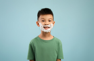 Little asian boy with shaving foam on face posing over blue background