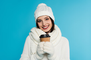 Positive woman in gloves and warm scarf holding paper cup isolated on blue