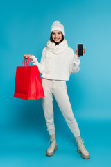 Smiling woman in knitted clothes showing smartphone with blank screen and shopping bags on blue background