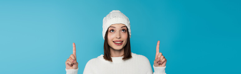 Brunette woman in knitted hat pointing with fingers isolated on blue, banner