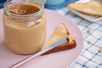 knife with creamy peanut butter on a slice of toast. A full jar of peanut butter and toaster bread on a plate close . Healthy breakfast concept. High quality photo