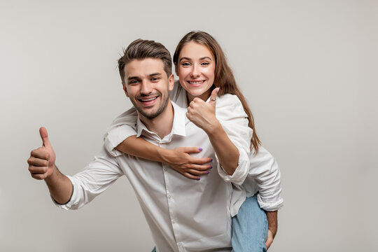 Image of excited couple having fun while man piggybacking joyful woman with thumb up isolated over white background