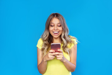 A young caucasian amazed joyful blonde woman with wavy hair in a yellow t-shirt smiles holding a mobile phone looking at the screen isolated on a color blue background