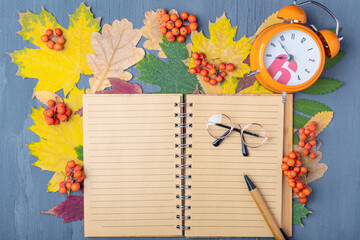 Orange alarm clock, blank lined craft notepad, pen and glasses on a background of autumn dry colorful leaves. Working day planning concept. Autumn time management.