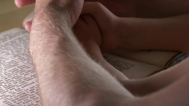Father overlays his hands with his son's in prayer over Bible scripture. Static shot.