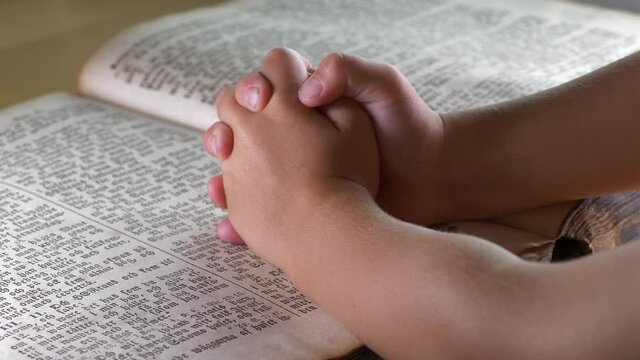 Young child clasps hands in prayer over bible as father shows him how. Static shot. Close up.