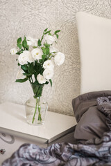 white flowers in a transparent glass vase stand on a bedside table by a bed with a large white headboard. bedroom interior. modern room. romantic. valentine's day 