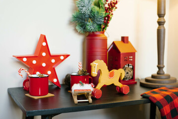 Christmas decor in the house: a wooden horse-gurney, a mug with cocoa, stars and a house on the dresser