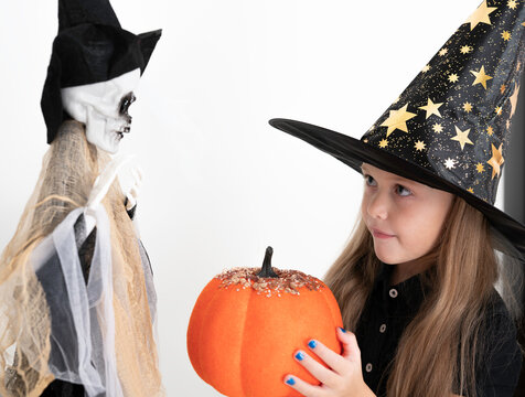 girl dressed as a witch, with a hat on her head, gives an orange pumpkin to a suspended skeleton. Halloween celebration, party