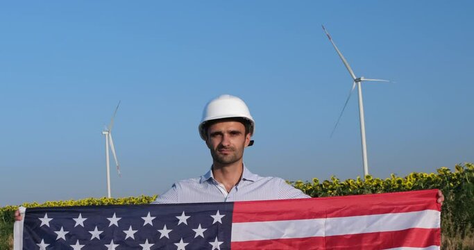 A man in a protective helmet holds the US flag in front of him against the backdrop of a field of sunflowers and wind turbines. USA Engineer for Clean Renewable Energy.