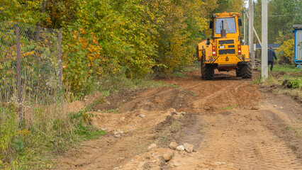 Rural road construction with grader in autumn daytime. The grader repairs the dirt road in the...