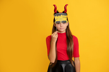 serious teen girl wearing imp horns holding party accessory on yellow background, halloween