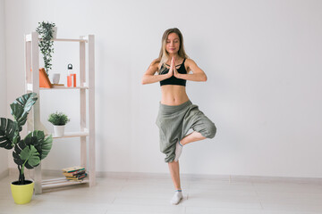 Woman practise yoga performs. Tree pose standing inside of cozy room with plants and greenery. Healthy lifestyle and mindfulness concept