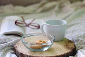 Bowl of cookies, cup, book and reading glasses on a bed. Selective focus.