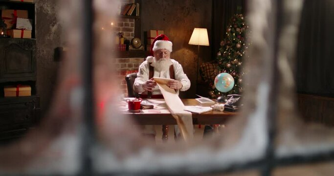Busy Santa Claus working at night, reading long wishlist, outside view through window. St Nicholas making up checklist of children wishes and presents for kids. Late work in evening on Christmas Eve.