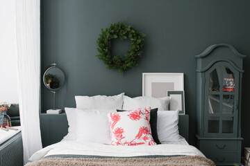 Pillows on the bed, above which a Christmas wreath hangs on a dark wall. Chest of drawers, mirror and mockups photo frames