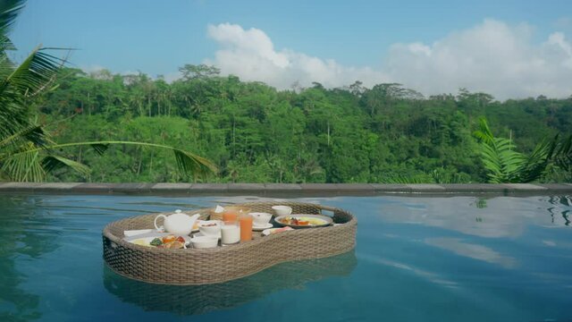 A basket with a morning breakfast floats in the view pool against the background of a tropical forest with palm trees against the background of a blue sky with clouds Luxury breakfast with fresh juice