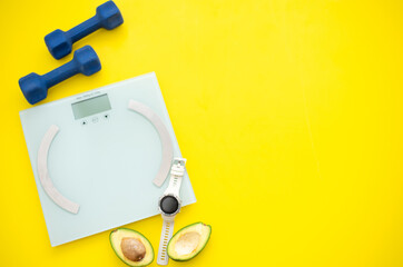 Cut avocado, weigher, dumbbells and sport watch on yellow background. Fruits contain many vitamins. healthy fruits:  good for body. food for diet. top view.  place for text.