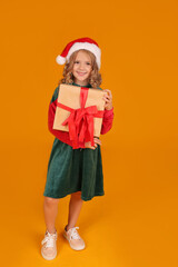 a beautiful blonde girl in a red and green Santa Claus hat dress holds a gift box on a yellow background
