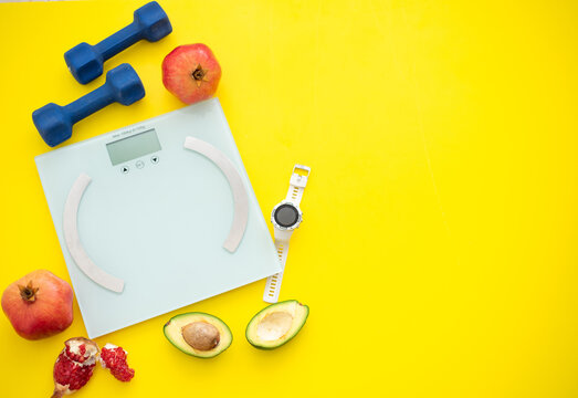 Three pomegranates, cut avocado, weigher, dumbbells and sport watch on yellow background. Fruits contain many vitamins. healthy fruits:  good for body. food for diet. top view. place for text 