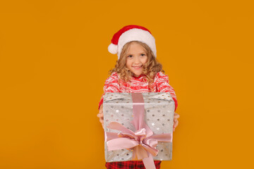 a little beautiful happy blonde girl in a Christmas jumper Santa Claus hat is holding a gift box on a yellow background