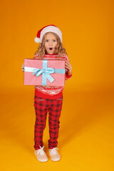 a little beautiful happy blonde girl in a New Year's jumper and plaid trousers with a Santa Claus hat is holding a gift box on a yellow background