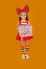 a little happy beautiful blonde girl with long hair with a red bow on her head in a colored blouse red skirt and colored socks on a yellow background holds a gift box in her hands