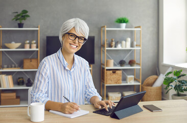 Portrait of senior business woman in glasses at table with coffee writing in paper notepad diary and working on modern smart tablet device PC to manage budget, do accounts, calculate money and taxes