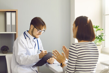 Obraz premium Male doctor writes prescription or makes notes in patient's card at medical examination. Doctor listens to woman who talks about health complaints sitting with her back to the camera.